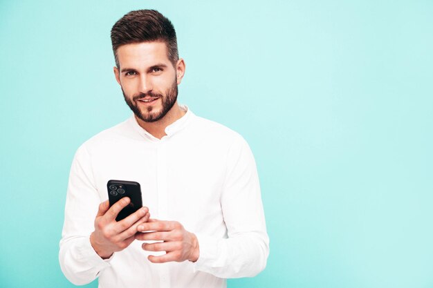 Bonito modelo sorridente Homem elegante sexy vestido de camisa e jeans Moda hipster masculino posando perto da parede azul no estúdio segurando o smartphone olhando para a tela do celular usando aplicativos