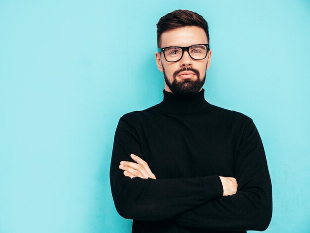 Bonito modelo sorridente Sexy homem elegante vestido com suéter preto de gola alta e jeans Moda hipster masculino posando perto da parede azul no estúdio Isolado em óculos ou óculos Braços cruzados