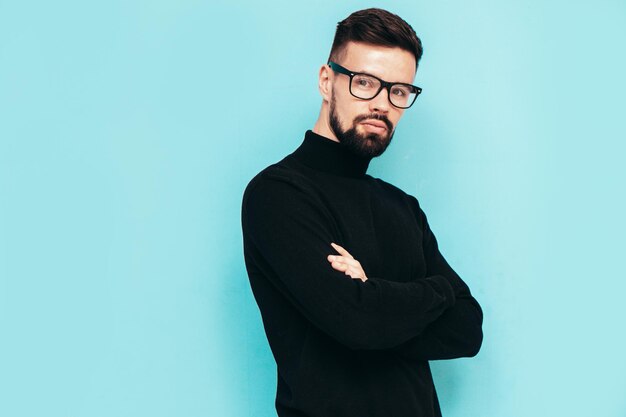 Bonito modelo sorridente Sexy homem elegante vestido com suéter preto de gola alta e jeans Moda hipster masculino posando perto da parede azul no estúdio Isolado em óculos ou óculos Braços cruzados
