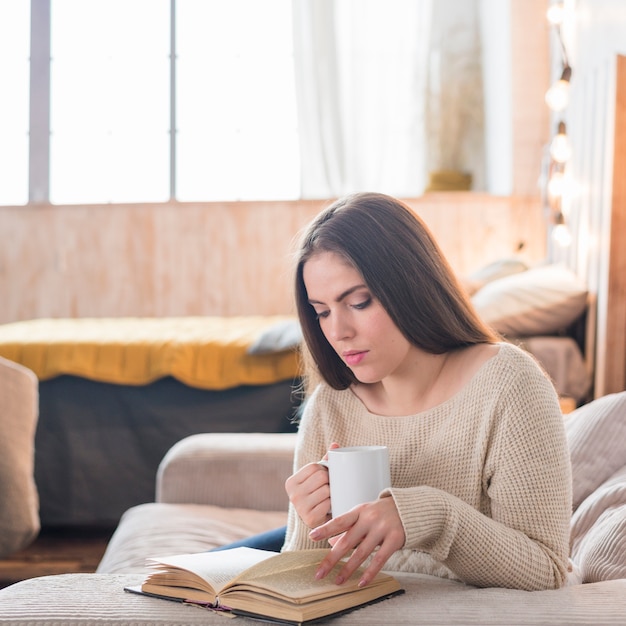 Bonito, mulher jovem, segurando, xícara café, leitura, livro