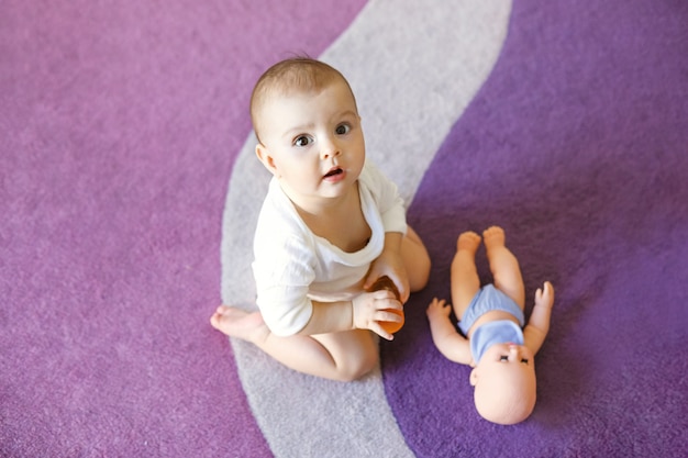 Bonito pequeno bebê mulher sentada no tapete roxo com boneca.