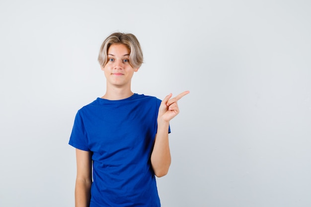 Bonito rapaz adolescente apontando para o canto superior direito em t-shirt azul e olhando esperançoso, vista frontal.