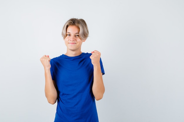 Bonito rapaz adolescente numa t-shirt azul, mostrando o gesto do vencedor e parecendo feliz, vista frontal.