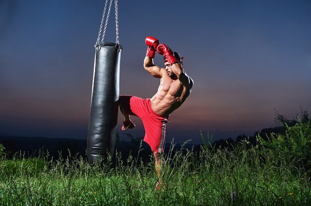 Bonito sem camisa muscular jovem kick boxer malhando com um saco de pancadas ao ar livre copyspace belo pôr do sol no fundo natureza estilo de vida esportes atleta ativo treinamento de masculinidade atlética.