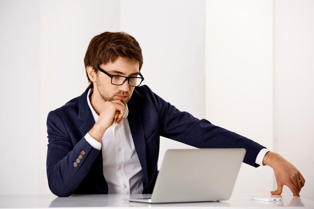Bonito, sério, determinado empresário usa óculos, lendo o relatório ou estudando gráficos na tela do laptop, trabalhando no escritório