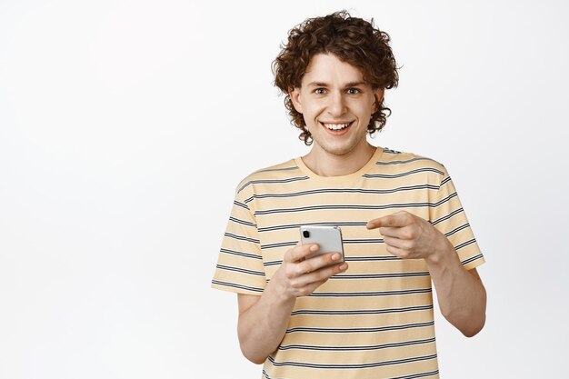 Bonito sorridente adolescente apontando para seu celular falando sobre app ou loja online sobre fundo branco