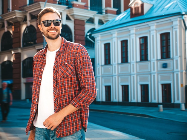 Foto grátis bonito sorridente elegante hipster lambersexual modelo. homem moderno vestido com camisa quadriculada vermelha. moda masculina posando perto de arranha-céus no fundo da rua em óculos de sol. ao ar livre ao pôr do sol