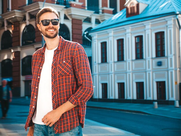 Bonito sorridente elegante hipster lambersexual modelo. Homem moderno vestido com camisa quadriculada vermelha. Moda masculina posando perto de arranha-céus no fundo da rua em óculos de sol. Ao ar livre ao pôr do sol