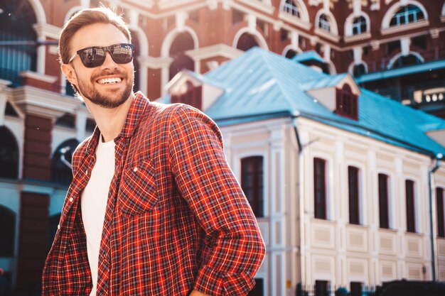Bonito sorridente elegante hipster lambersexual modelo. Homem moderno vestido com camisa quadriculada vermelha. Moda masculina posando perto de arranha-céus no fundo da rua em óculos de sol. Ao ar livre ao pôr do sol