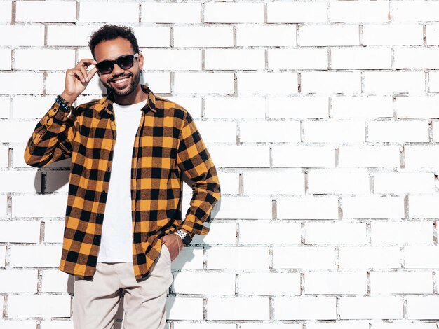 Bonito sorridente modelo hipster Homem com barba sexy vestido com camisa amarela de verão e roupas jeans Moda masculina posando perto da parede de tijolos brancos no estúdio em dia ensolarado isolado em óculos de sol