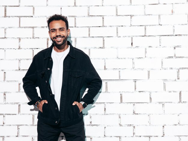 Bonito sorridente modelo hipster Homem com barba sexy vestido com camisa preta de verão e roupas jeans Moda masculina posando perto da parede de tijolos brancos no estúdio em dia ensolarado isolado