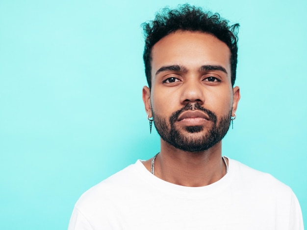 Bonito sorridente modelo hipster Homem com barba sexy vestido com camiseta branca de verão e roupas jeans Moda masculina posando perto da parede azul no estúdio isolado