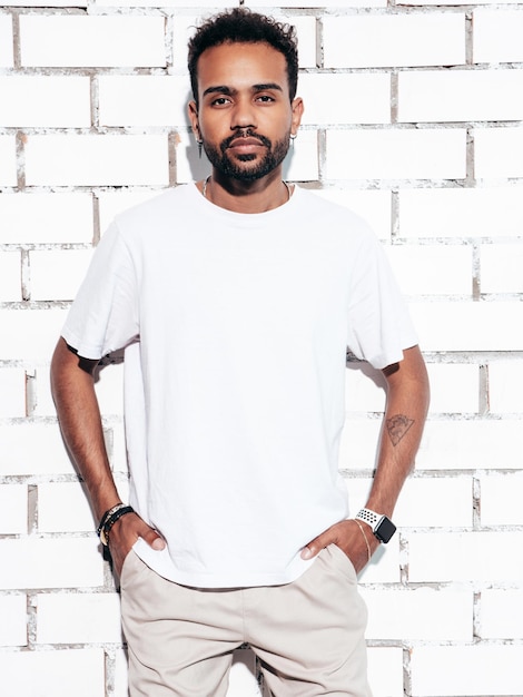 Bonito sorridente modelo hipster Homem com barba sexy vestido com roupas jeans e camiseta branca de verão Moda masculina posando perto da parede de tijolos brancos no estúdio em dia ensolarado isolado