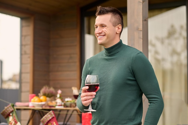 Bons momentos. feliz jovem adulto atraente de suéter verde com copo de vinho em pé perto da varanda festivamente decorada da casa sorrindo olhando de lado na tarde