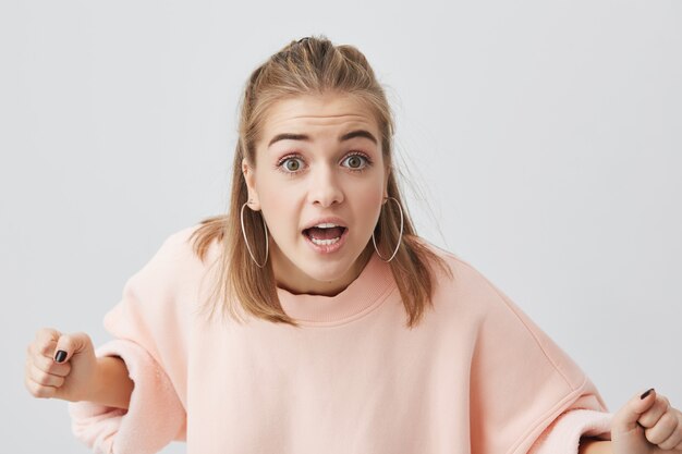 Brava fêmea jovem caucasiana com pele limpa saudável, vestindo blusa rosa posando isolado, estalando os olhos, prontos para discutir. Emoções negativas, insatisfação, agressão.