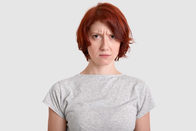 Brava mulher descontente com cabelo vermelho, expressa sentimentos negativos, carranca o rosto em descontentamento, veste camiseta casual, isolada sobre parede branca, parece descontente. Conceito de pessoas e emoções