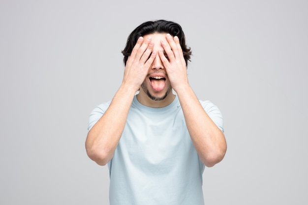 Brincalhão jovem bonito fechando os olhos com as mãos mostrando a língua isolada no fundo branco