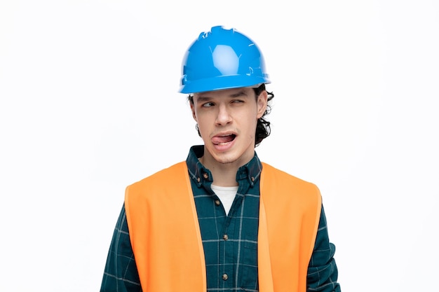 Brincalhão jovem engenheiro masculino usando capacete de segurança e colete de segurança olhando para o lado mostrando a língua isolada no fundo branco