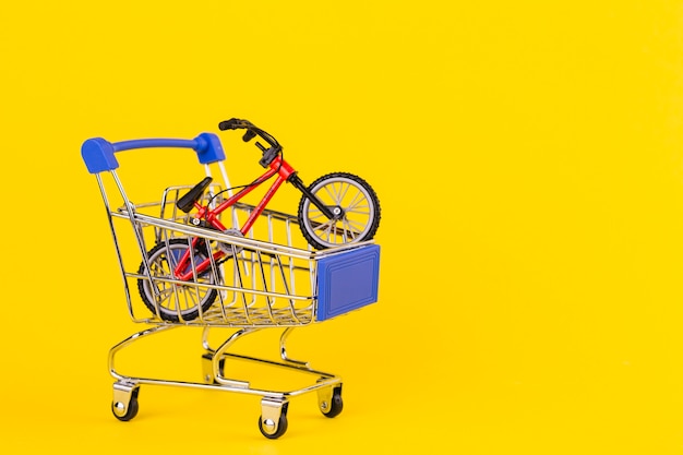 Brinquedo pequeno da bicicleta no carrinho de compras contra o fundo amarelo