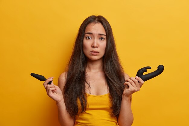 Brinquedos sexuais, aparelhos de masturbação. Mulher asiática preocupada segura vibrador de silicone e plug anal, se prepara para jogos sexuais íntimos, se dá prazer com o vibrador, fica de pé contra a parede amarela. Intimidade