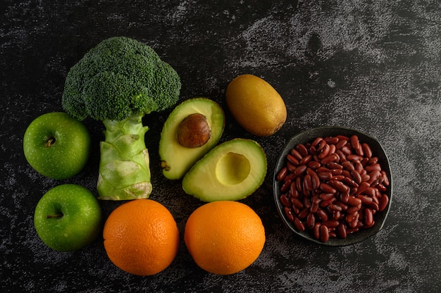 Brócolis, maçã, laranja, kiwi, feijão vermelho e abacate em um piso de cimento preto.