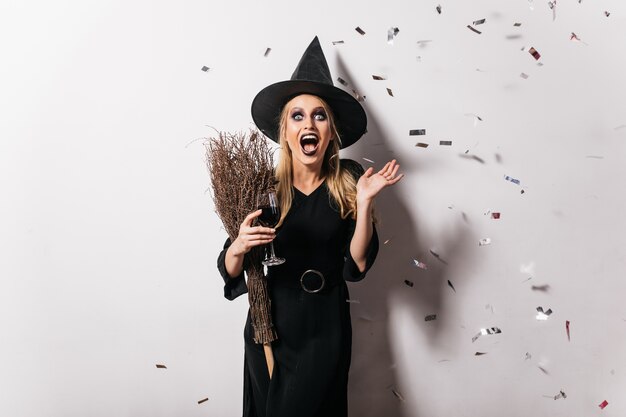 Bruxa bonita espantada com chapéu, bebendo vinho. Mulher loira simpática de vestido preto relaxante no halloween.