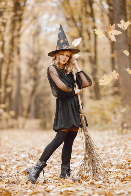 Bruxa loira jovem na floresta no Halloween. Garota usando vestido preto e chapéu de cone. Bruxa segurando uma vassoura.