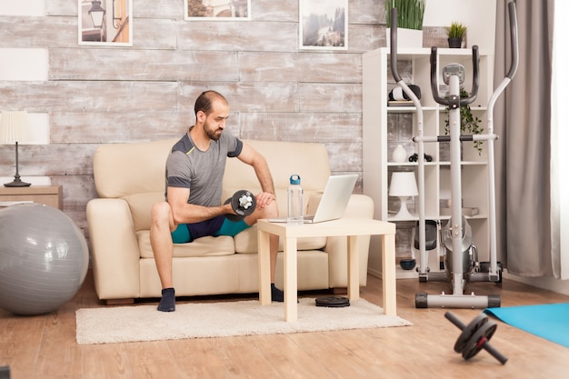 Cabe o homem no sofá, treinando bíceps com halteres, assistindo ao treino online em tempo de auto-isolamento.