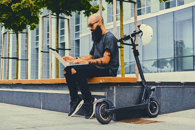 Cabeça raspada tatuada, homem barbudo usando um laptop sobre o fundo do edifício moderno após o passeio de scooter elétrico.