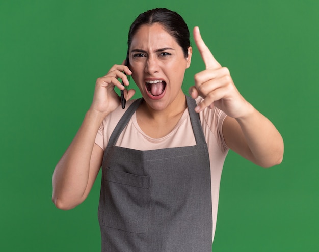 Cabeleireira jovem bonita com raiva de avental falando no celular, gritando com uma expressão agressiva, mostrando o dedo indicador em pé sobre a parede verde