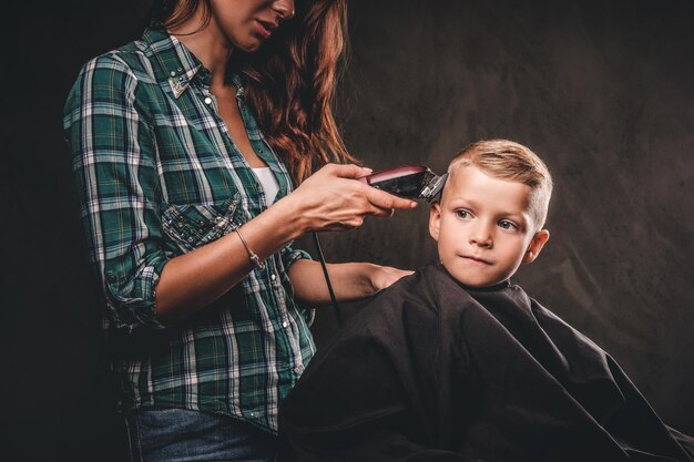 Cabeleireiro de crianças com o aparador está cortando garotinho contra um fundo escuro. Menino bonito da pré-escola cortando o cabelo.