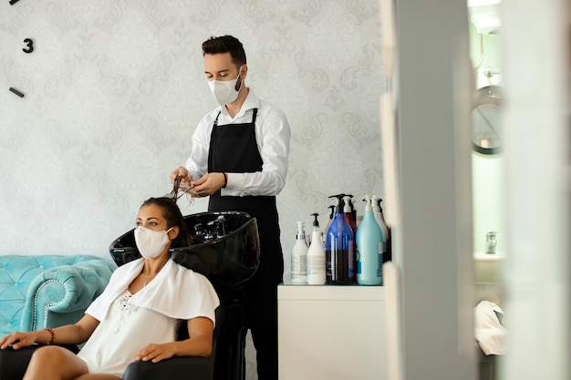 Cabeleireiro e seu cliente usando máscara facial protetora durante o tratamento capilar no salão