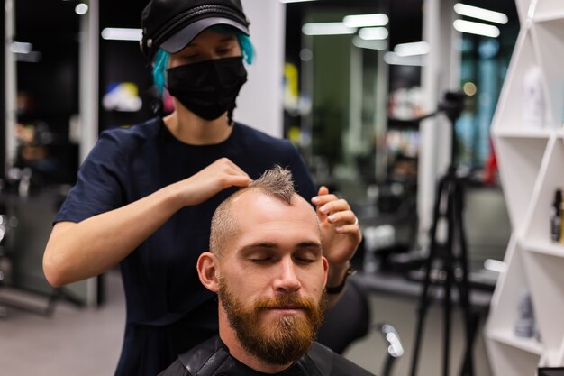 Cabeleireiro profissional usando máscara protetora, faz corte de cabelo para homem barbudo brutal em salão de beleza