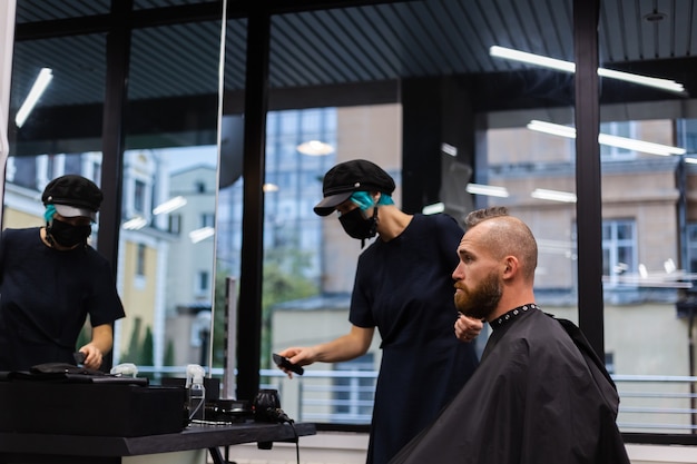 Cabeleireiro profissional usando máscara protetora, faz corte de cabelo para homem barbudo brutal em salão de beleza