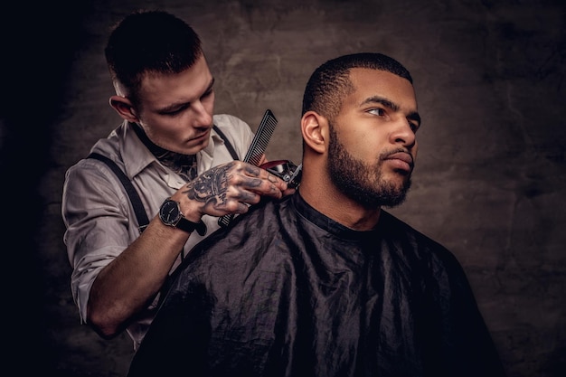 Cabeleireiro tatuado profissional antiquado faz um corte de cabelo para um cliente afro-americano, usando um aparador e um pente. isolado em plano de fundo texturizado escuro.