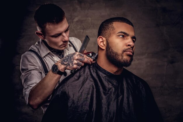 Cabeleireiro tatuado profissional antiquado faz um corte de cabelo para um cliente afro-americano, usando um aparador e um pente. Isolado em plano de fundo texturizado escuro.