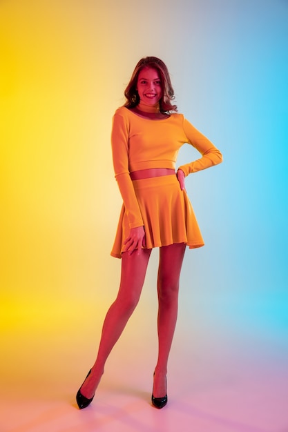Cabelo longo. Linda garota sedutora num vestido elegante em fundo gradiente amarelo-azul com luz de néon.