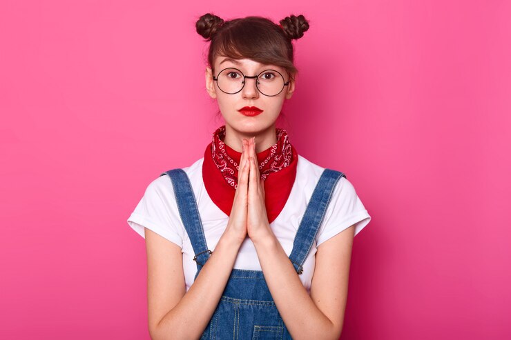 Foto grátis cabelos castanhos olhos femininos jovem em pé isolado sobre rosa, rezando com juntar as mãos, olhando diretamente para a câmera, tendo cachos e lábios coloridos brilhantes.