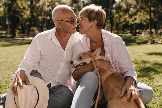 Cabelos curtos em blusa rosa listrada, posando com pequeno corgi e beijando o homem com bigode em camisa branca e jeans no parque.