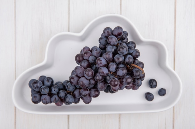 Foto grátis cacho de uvas pretas em uma placa em forma de nuvem em fundo branco vista superior