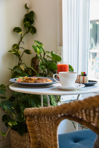 Cadeira de vime, perto da mesa redonda branca com uma xícara de café; café da manhã; smoothie e cheesecake