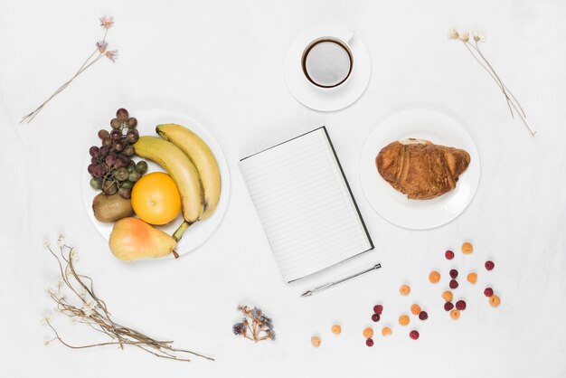 Caderno; caneta; croissant; frutas; café e flores secas em pano de fundo branco