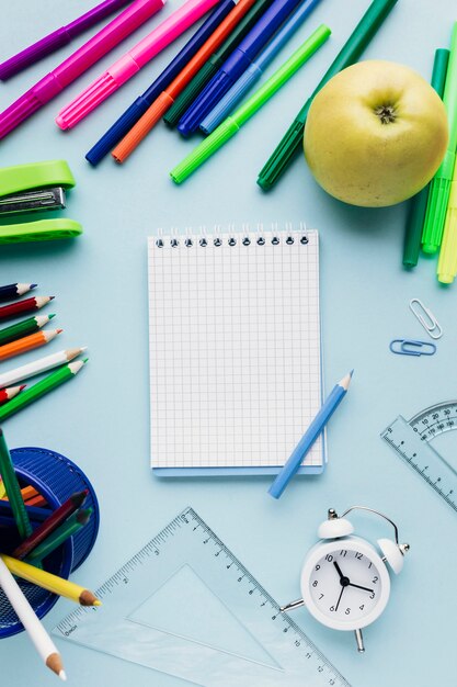 Caderno claro rodeado de papelaria brilhante, relógio e apple em fundo azul