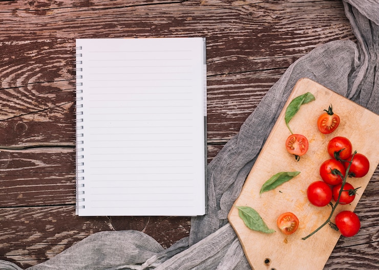 Foto grátis caderno espiral de linha única; tomate cereja e manjericão em cortar a placa sobre a mesa de madeira