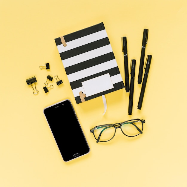Caderno fechado com canetas de feltro; clipes de papel de bulldog; óculos e celular em fundo amarelo
