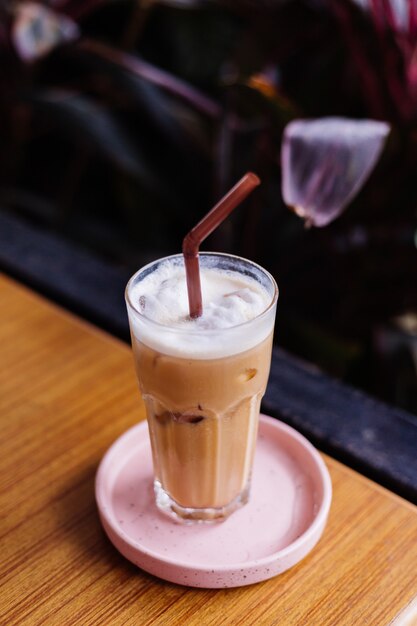 Café com leite gelado em vidro em um suporte rosa em uma mesa de madeira em arbustos verdes de café de verão