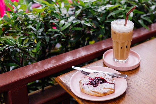 Café com leite gelado em vidro em um suporte rosa na mesa de madeira e torta de cranberry em arbustos verdes de café de verão