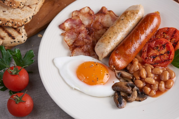 Café da manhã inglês completo tradicional com ovos fritos, salsichas, feijão, cogumelos, tomates grelhados e bacon em um prato, torradas, manteiga e geléia em uma placa de madeira