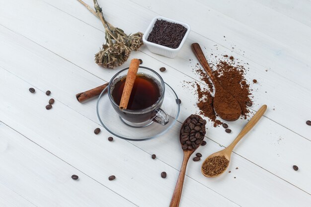 Café em uma xícara com café moído, grãos de café, paus de canela e ervas secas, vista de alto ângulo em um fundo de madeira