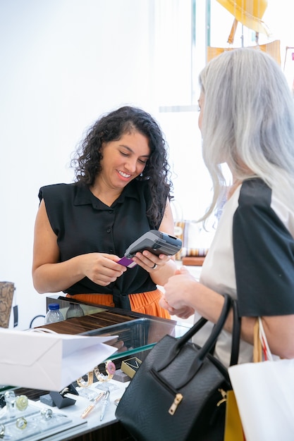 Caixa da loja ou vendedor positivo falando com o cliente e operando o processo de pagamento com terminal POS e cartão de crédito. Tiro médio. Conceito de compra ou compra